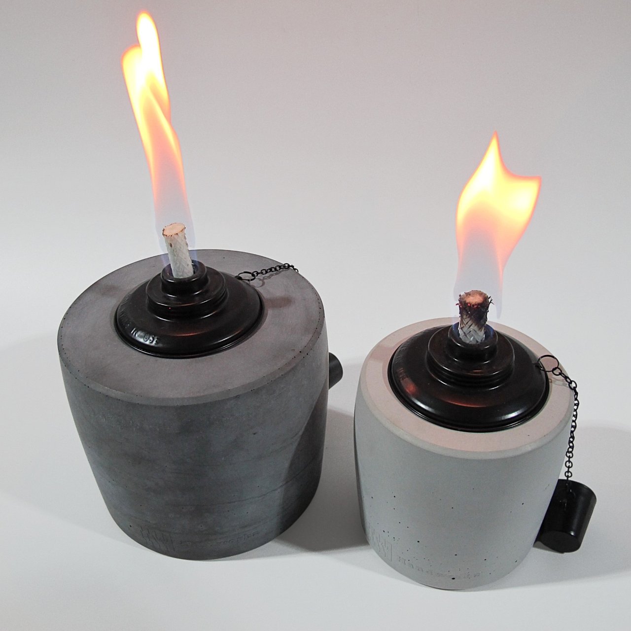 Links im Bild ist ein großes Betonfeuer in Anthrazit mit einer lang gezogenen Flame in schönem gelb/rot. Rechts daneben steht der kleine Bruder in der Farbe Grau, die Flame ist etwas breiter und scheint ebenso im schönen gelb/rot. Am unteren Rand der Gartenfackeln ist leicht das Branding der Marke Handworkz zu sehen, es ist in einem Guss mit dem Beton und somit sehr dezent und stilvoll.