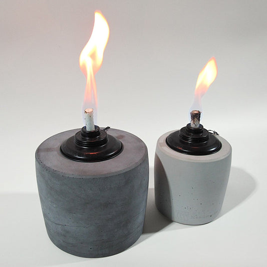 Wie beim vorherigen Bild ist Links im Bild ein großes Betonfeuer in Anthrazit mit einer lang gezogenen Flame in schönem gelb/rot, diesmal ist rechts daneben steht kleine Bruder in der Farbe Grau etwas nach hinten versetzt, die Flame ist etwas kleiner als die der großen Öllampe und scheint ebenso im schönen gelb/rot. Am unteren Rand der Gartenfackeln ist leicht das Branding der Marke Handworkz zu sehen, es ist in einem Guss mit dem Beton und somit sehr dezent und stilvoll.