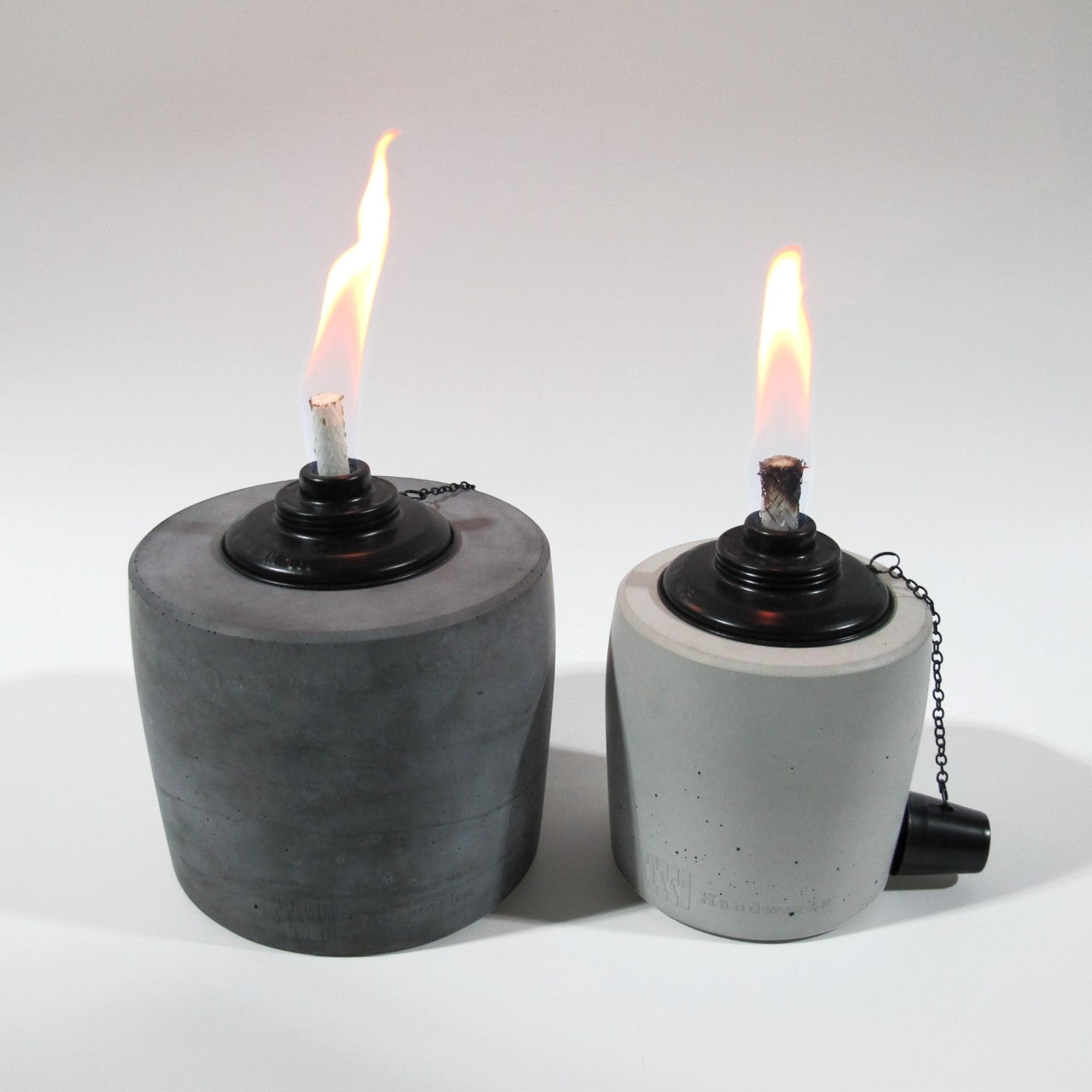 Links im Bild ein großes Betonfeuer in Anthrazit mit einer lang gezogenen Flame in schönem gelb/rot, rechts daneben steht der kleine Bruder in der Farbe Grau perfekt auf einer Höhe um den Größenunterschied der Betonfackeln schön zu sehen, die Flame ist schmal und scheint ebenso im schönen gelb/rot. Am unteren Rand der Gartenfackeln ist leicht das Branding der Marke Handworkz zu sehen, es ist in einem Guss mit dem Beton und somit sehr dezent und stilvoll.