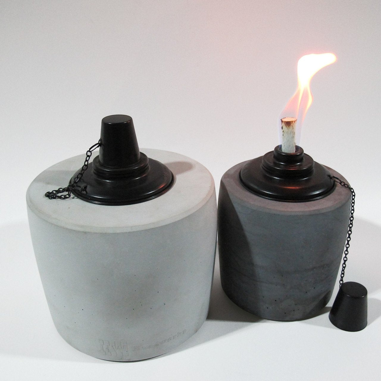 Links im Bild ist eine große Graue Öllampe in der Farbe Grau zu sehen, geprägt von dem Logo der Marke Handworkz und mit geschlossenem Löschdeckel. Rechts daneben steht die kleine Version der Fackel in Anthrazit mit gezündeter Flame, als Brennstoff wurde Bioethanol verwendet.